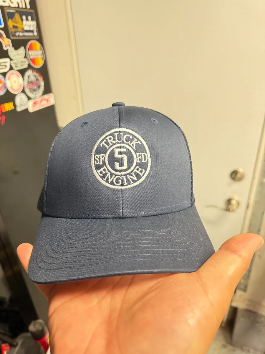 Grey cap embroidered with an SFFD Truck 5 Engine roundel, held up in the firehouse