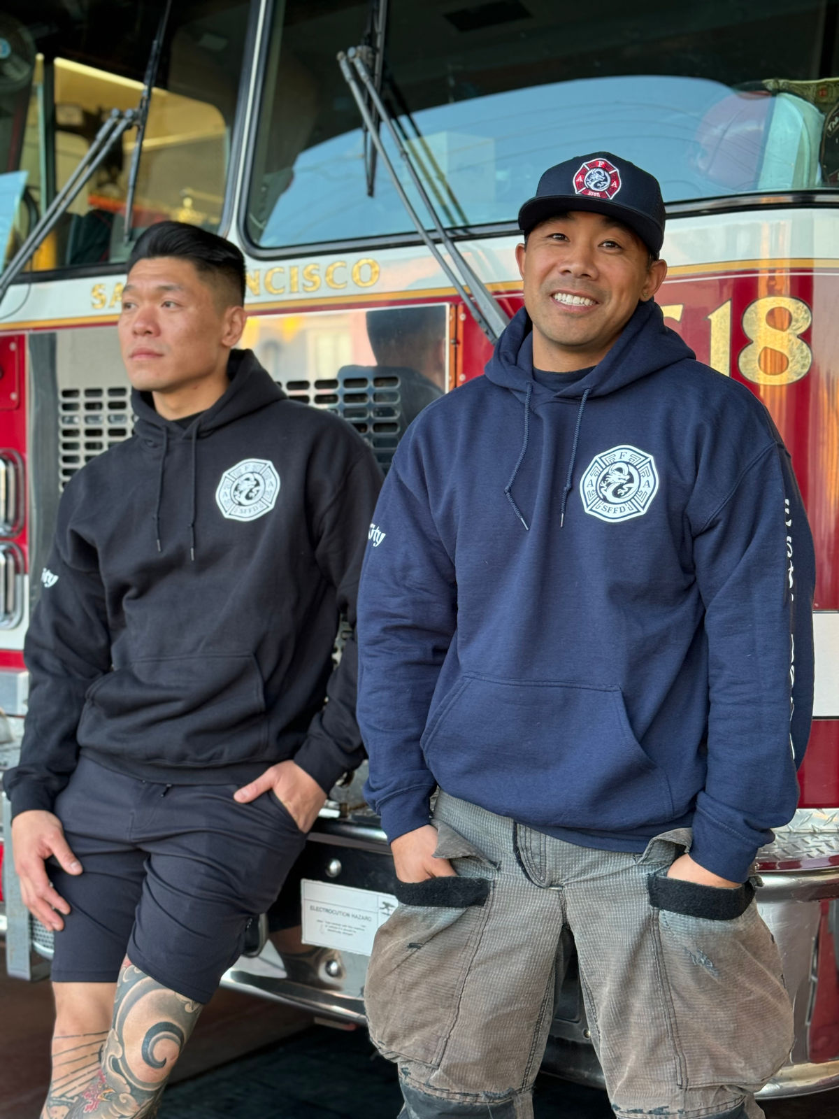 Two firefighters in station hoodies leaning against the front of their rig