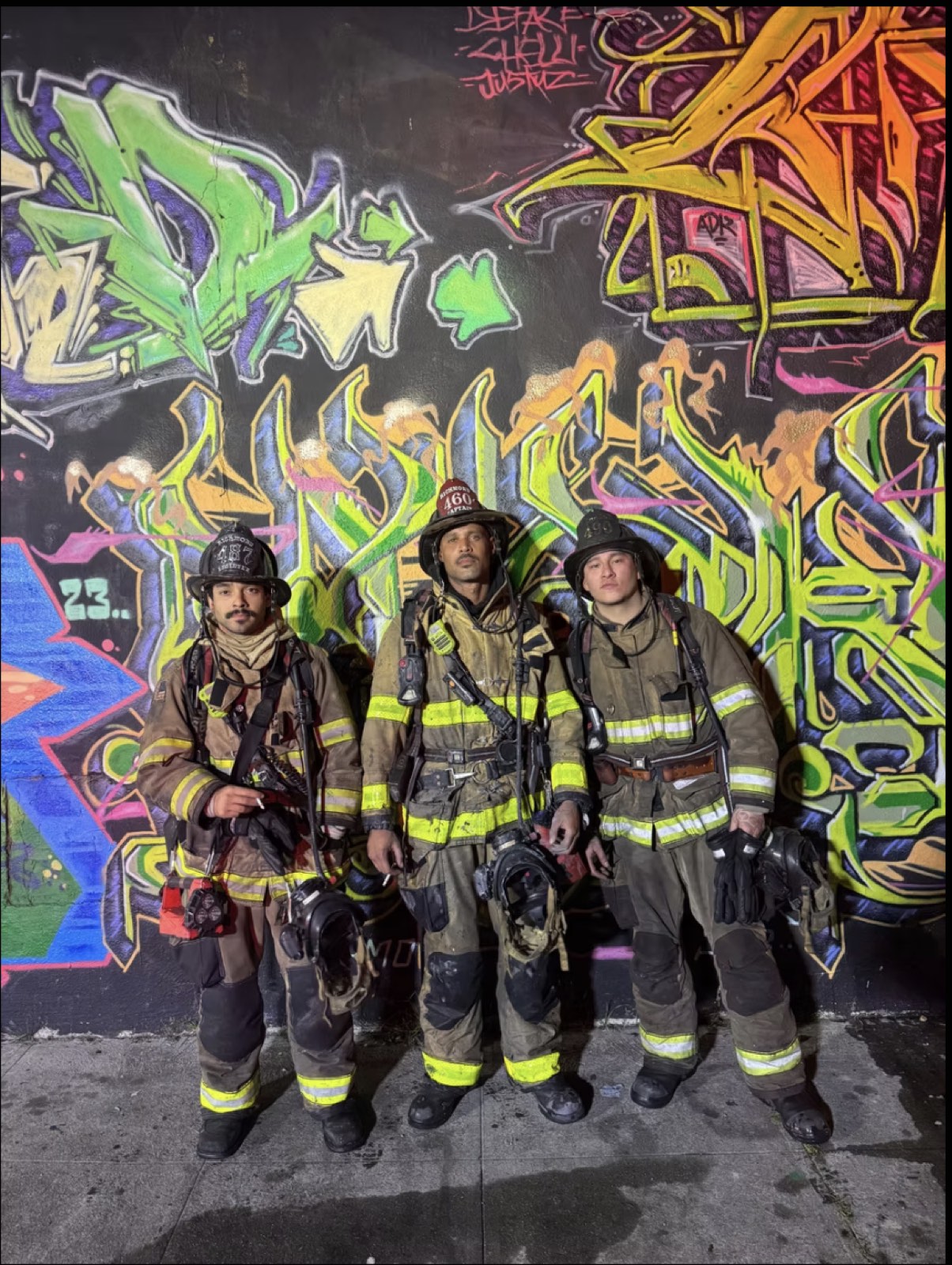 Three firefighters in turnouts standing against a wall of graffiti